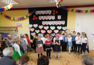 Przedszkolaki ustawione w półkolu, na środku stojące dwie dziewczynki i chłopiec śpiewające piosenkę