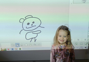 Dziewczynka z młodszej grupy prezentuje narysowanego misia na tablicy interaktywnej