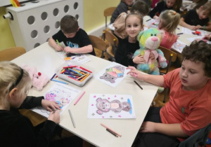 Przedszkolaki siedzą przy stoliku i kolorują obrazki misia.