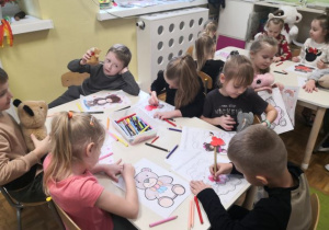 Dzieci siedzą przy stoliku i kolorują obrazki misia.
