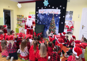 Przedszkolaki ubrane na czerwono witają Mikołaja. W tle piękna zimowo-świąteczna dekracja.
