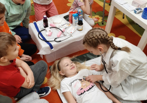 Dzieci z grupy 5,6 latków podczas zabawy dramowej w lekarza. dziewczynka w białym fartuchu osłuchuje leżącą na leżance koleżankę.