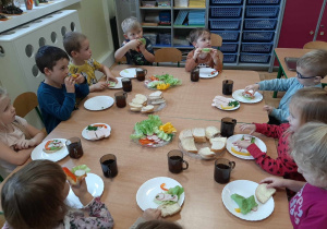 Dzieci z grupy 3,4,5 latków siedzą przy stole, na którym są zdrowe produkty. Przygotowują zdrowe i kolorowe kanapki.