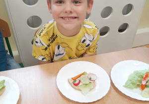 Chłopiec z grupy 3,4,5 latków prezentuje przy stoliku przygotowaną zdrową kanapkę.