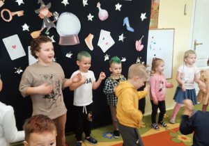 Grupa dzieci tańczy podczas andrzejkowej zabawy. Prezentują swoje dziwne fryzury. W tle andrzejkowa dekoracja.