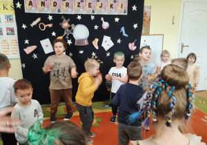 Grupa dzieci tańczy podczas andrzejkowej zabawy. Prezentują swoje dziwne fryzury. W tle andrzejkowa dekoracja. Inne ujęcie.