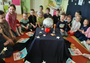 Przedszkolaki siedzą na dywanie wokół magicznego stolika, na którym stoi srebrna kula, świeci elektryczna świeczka.