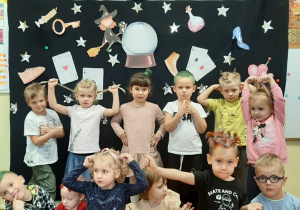 Dzieci z grupy 3,4,5 latków pozują do fotografii na tle andrzejkowej dekoracji. Robią śmieszne miny, prezentują fryzury.