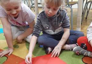 Dwie dziewczynki siedzą na dywanie. Jedna z nich przebija papierowe serce pinezką.