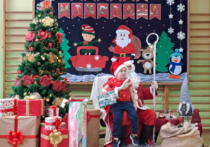 Mikołaj z przedszkolakiem na kolanach. W tle dekoracja świąteczna.