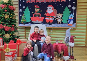 Dzieci siedzą w fotelu Mikołaja. W tle dekoracja świąteczna.