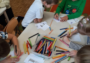 Dzieci siedzą przy stoliku rysują na kartkach wymarzone prezenty.