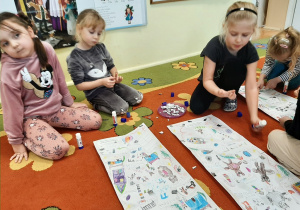 Kilka dziewczynek siedzi na dywanie dokleja kolorowe i błyszczące elementy ozdobne na list do Mikołaja.