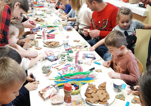 Dzieci i rodzice z grupy 5,6 latków siedzą przy długim stole, dekorują pierniki.