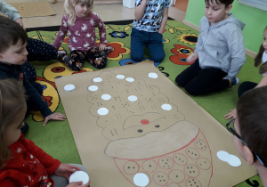 Dzieci na dywanie podczas zabawy wypełniają sylwetę Mikołaja białymi kołami.