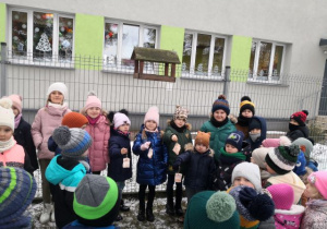 Przedszkolaki stoją koło karmnika, w środku stoją dzieci i trzymają słoninę dla ptaszków.