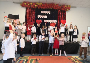 Przedszkolaki trzymają czerwone serca i śpiewają piosenkę.