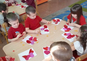 Przedszkolaki siedzą przy stolikach i wykonują pracę plastyczną z sercami