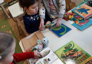 Dziewczynki siedzą przy stoliku, oglądają książki i ukladają puzzle z dinozaurami.