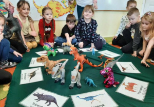Przedszkolaki siedzą w kole, w środku leżą obrazki z dinozaurami oraz figurki.