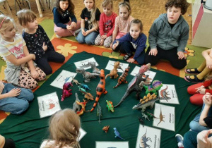 Przedszkolaki siedzą w kole, w środku leżą obrazki z dinozaurami oraz figurki, kolejne ujęcie.
