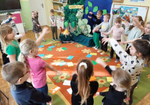 Dzieci stoją w dużym kole, uczestniczą w zabawie muzyczno-ruchowej. Na środku leżą ślady dinozaurów.