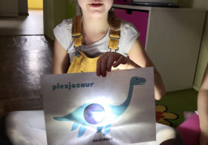 Dziewczynka podczas zabawy z latarką i obrazkiem dinozaura.