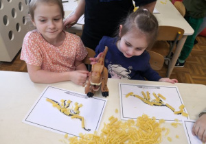 Dziewczynki siedzą przy stoliku i układają szkielety dinozaurów z makaronu.