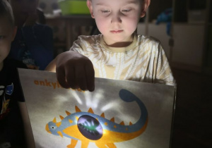 Chłopiec podczas zabawy z latarką i obrazkiem dinozaura.