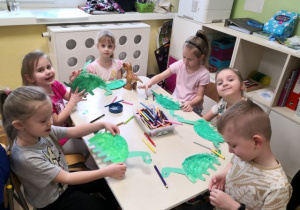 Dzieci siedzą przy stoliku i wykonują pracę plastyczną - dinozaury.