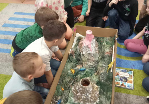 Dzieci siedzą na dywanie podczas eksperymentu. Na środku makieta z wulkanami i dinozaurami