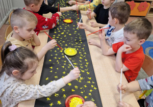 Dzieci siedzą przy długim stole i za pomocą słomki stemplują czarną rolkę papieru tworząc gwiazdy na niebie