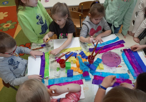 Dzieci siedzą przy stoliku, wykonują eksperyment "malująca bibuła" - zwilżają wodą paski bibuły ułożone na kartonie.