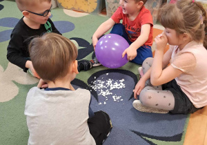 Doświadczenia z balonem. Na dywanie siedzą dzieci. Za pomocą balonu przyciągają kawałki papieru.