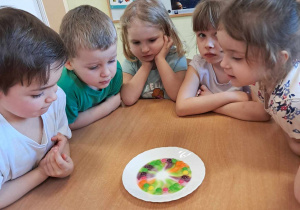 Dzieci przy stoliku obserwują jak na talerzyku rozpuszczają się kolorowe draże tworząc tęczowe pasy.