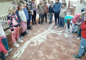 Przedszkolaki w kole. W środku ułożony z tektury szkielet dinozaura.