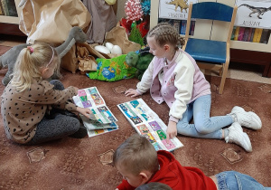 W bibliotece. Dzieci siedzą na dywanie i oglądają książki. W tle biblioteczny kącik dinozaurów.