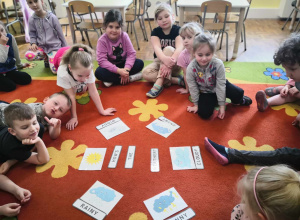 Dzieci siedzą w kole na dywanie. Na środku symbole ze zjawiskami atmosferycznymi. Dzieci uczestniczą w zabawie w języku angielskim.