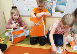 Przedszkolaki siedzą na dywanie podczas zabawy "Marcowy garnek". W wykonanym papierowym garnku umieszczają symbole pogody.