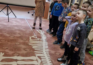 Dzieci stoją obok ułożonego na dywanie szkieletu dinozaura