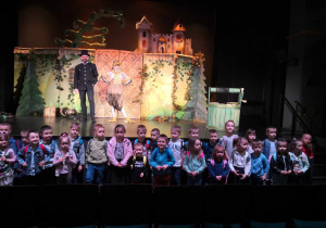 Dzieci z przedszkola stoją przed sceną i pozują do zdjęcia grupowego.