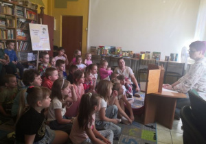 Dzieci siedzą w bibliotece na dywanie i słuchają legendy czytanej przez Panią Bibliotekarkę.