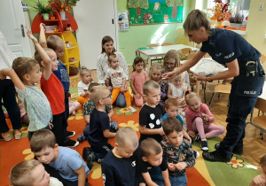 Pani policjantka rozdaje dzieciom siedzącym na dywanie odblaskowe breloczki.