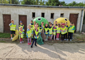 Dzieci z grupy starszaków pozują do zdjęcia z workami na śmieci i rękawicami. W tle kosze na śmieci na terenie przedzszkola.