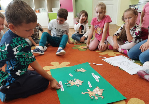 Dzieci siedzą na dywanie, chętne dziecko przykleja elementy na plakacie.