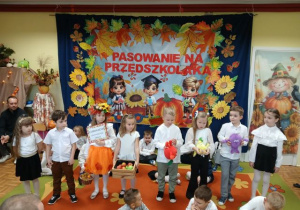 Przedszkolaki podczas recytacji wierszy. Dziewczynka przebrana za panią jesień trzyma zaproszenie i kosz z owocami. Dzieci tzrymają skrzynkę z owocami i pluszowe jabłko, gruszkę i śliwkę.