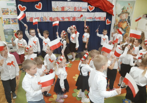 Dzieci maszerują w kole wymachując flagami do piosenki. W tle dekoracja tematyczna.