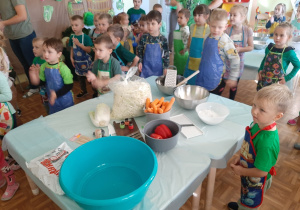 Dzieci stoją i wykonują ruchy do piosenki o kiszeniu kapusty. na pierwsym planie stół z produktami do kiszenia kapusty.