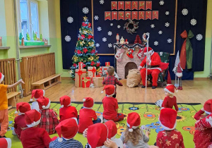 Dzieci siedzą na dywanie. W tle Mikołaj siedzi na fotelu.