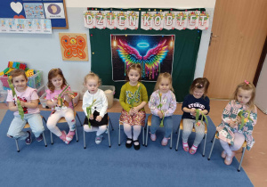 Dziewczynki z gr 3-4 latków siedzą z kwiatami na tle dekoracji